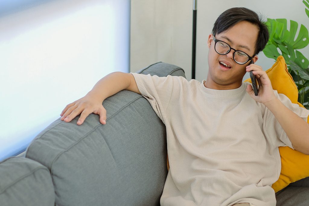 Cheerful Young Man with Down Syndrome Chatting on Smartphone at Home, Enjoying Leisure Time